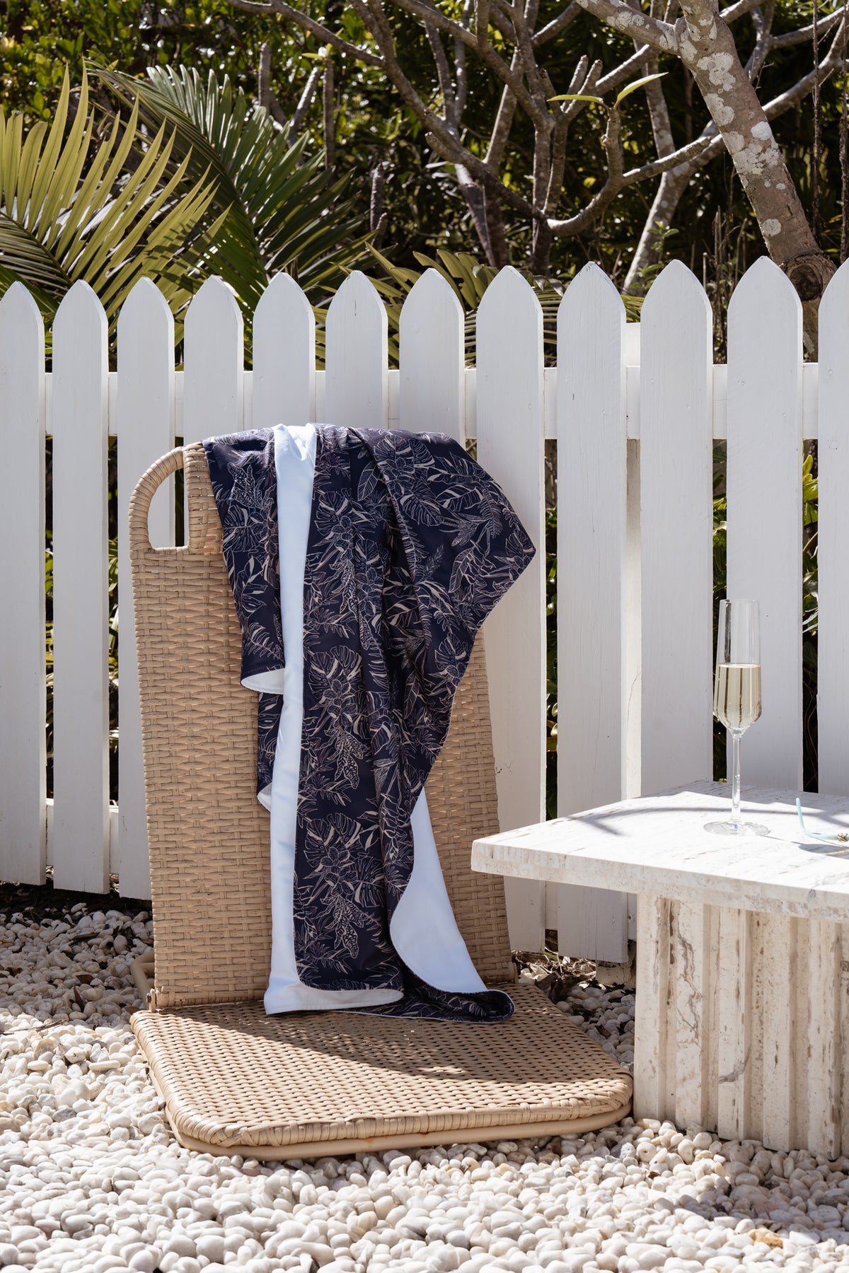 Neren Towel in Midnight Tropics hanging over a white picket fence beside a woven beach bag